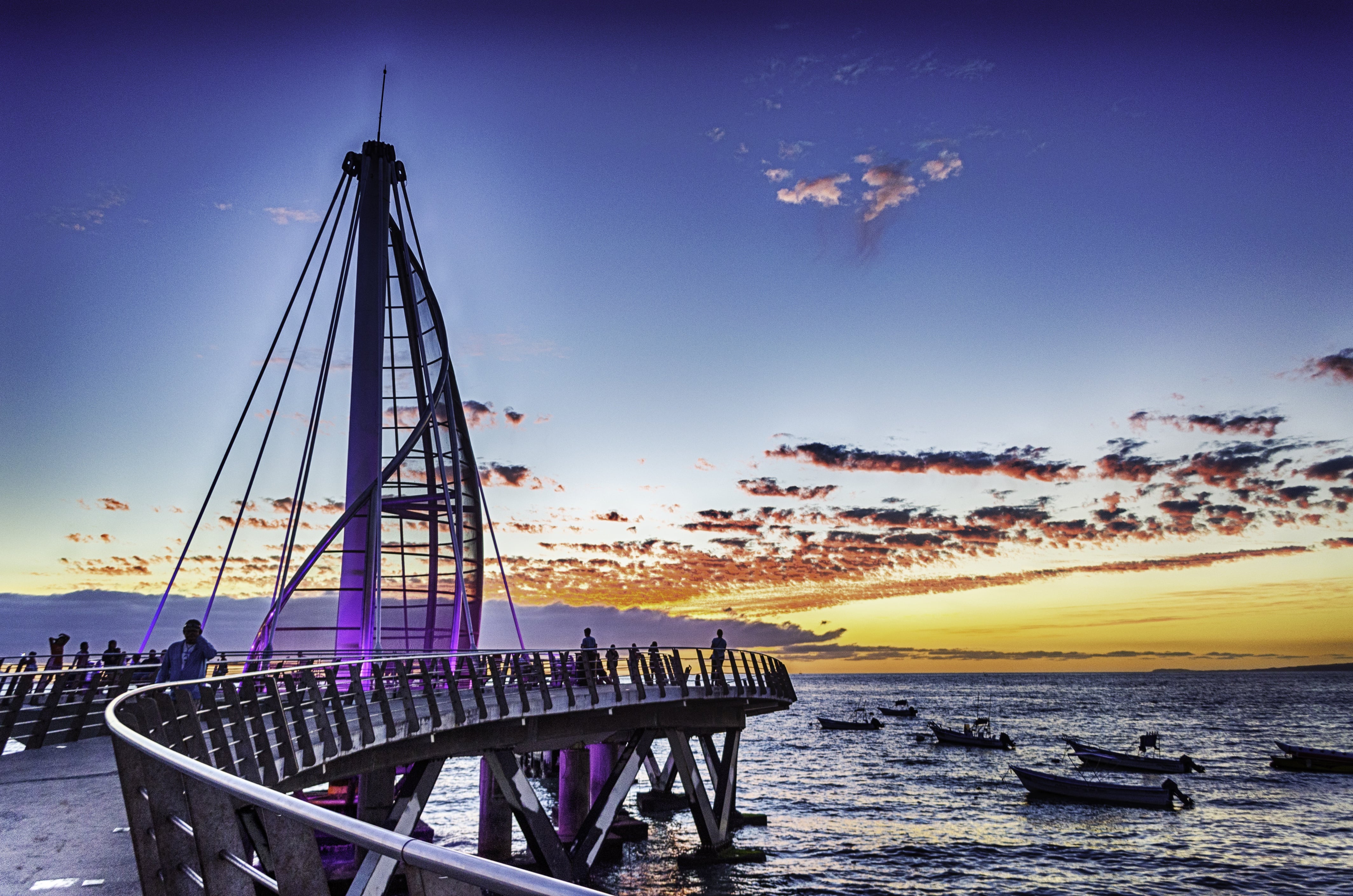 A picture of puerto vallartas boardwalk with muelle los muertos. Tall spiral with lights and a pear that extendes from to shore to wrap around it with boats around the dock.